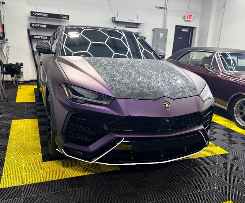 Lamborghini Urus in a satin purple colour-change wrap sitting inside the shop bay, hood panel still covered mid-job, film boxes racked on the wall behind