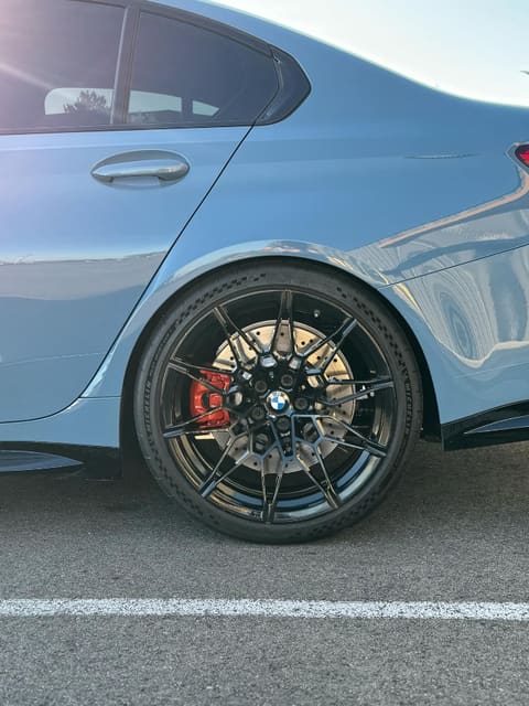 Rear wheel of a pale blue BMW M3: gloss black multi-spoke wheel with red brake calipers visible behind the spokes
