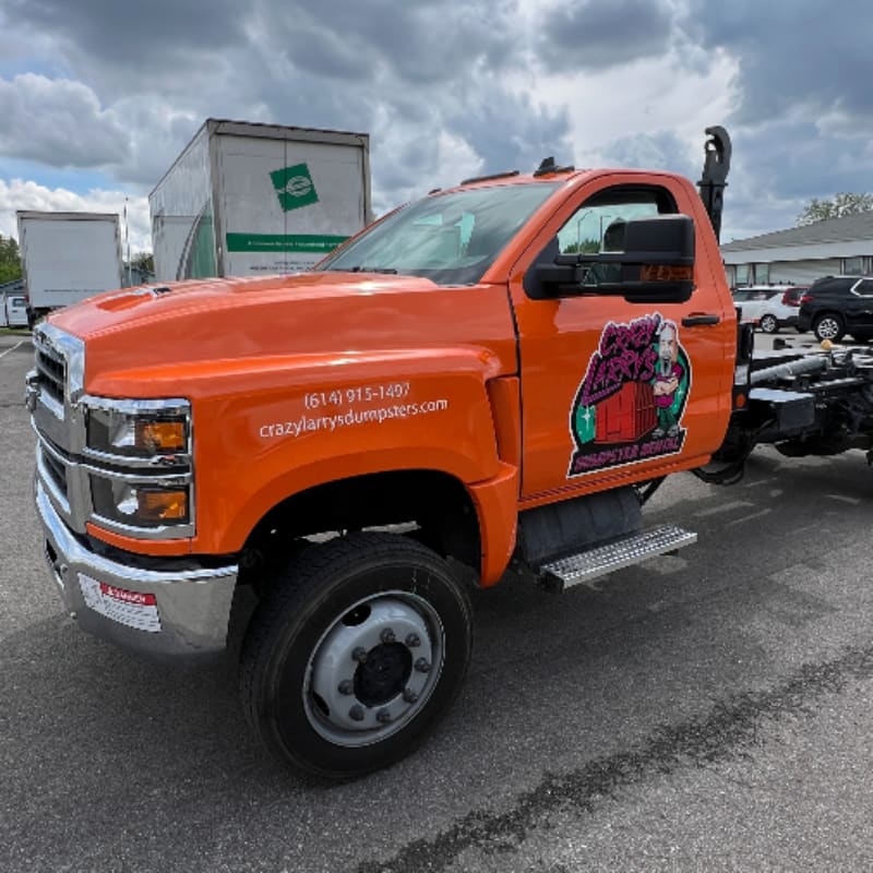Orange Chevrolet Silverado hooklift dumpster truck with fresh vinyl lettering, a cartoon dumpster logo and a 614 phone number on the door