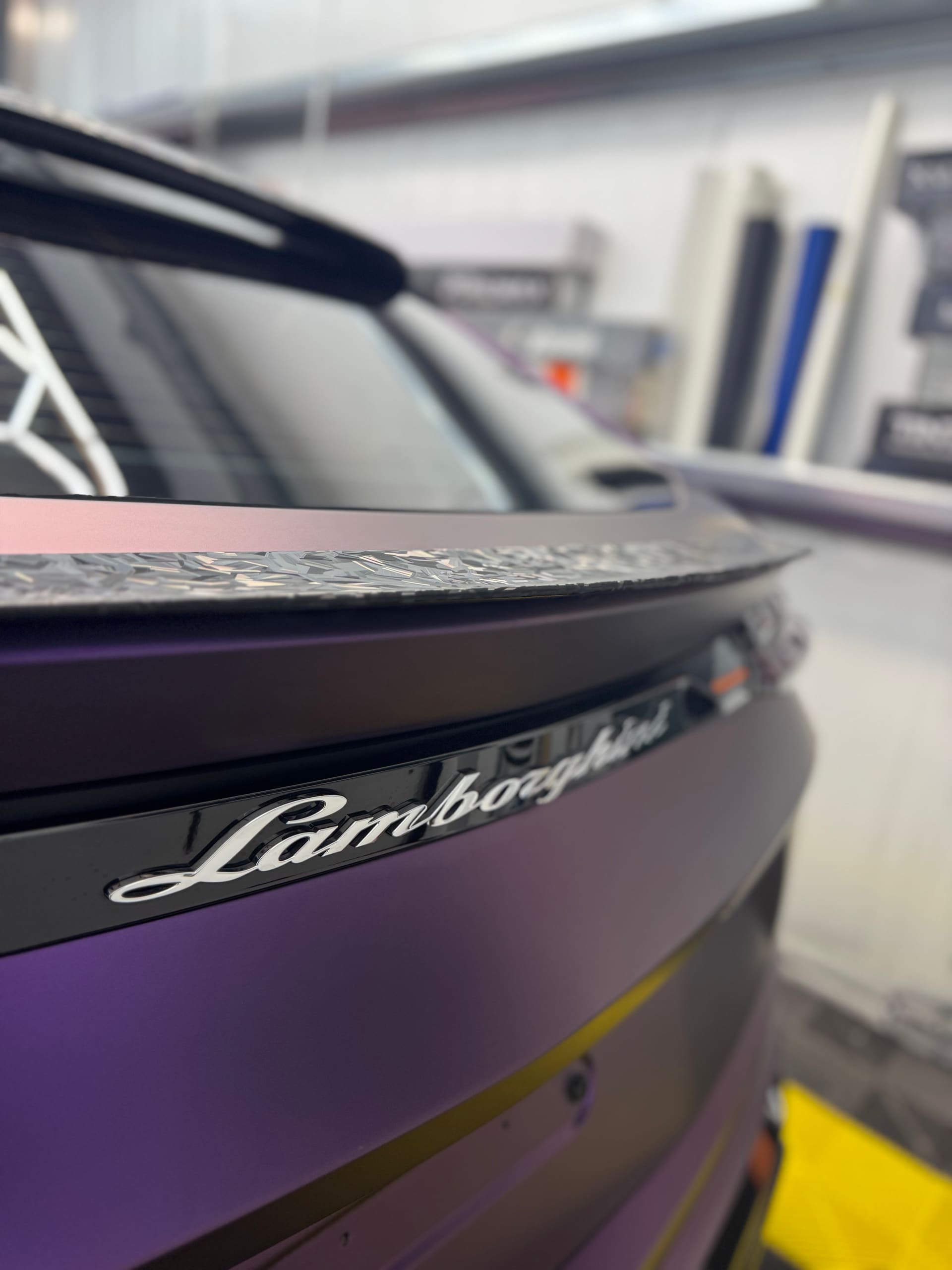 Close-up of the Lamborghini badge on the rear of a satin purple Urus, forged carbon spoiler in frame