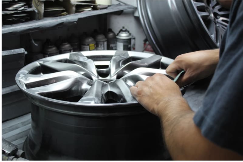Two hands steadying a freshly refinished silver alloy wheel on a workbench, a shelf of aerosols and bottles behind