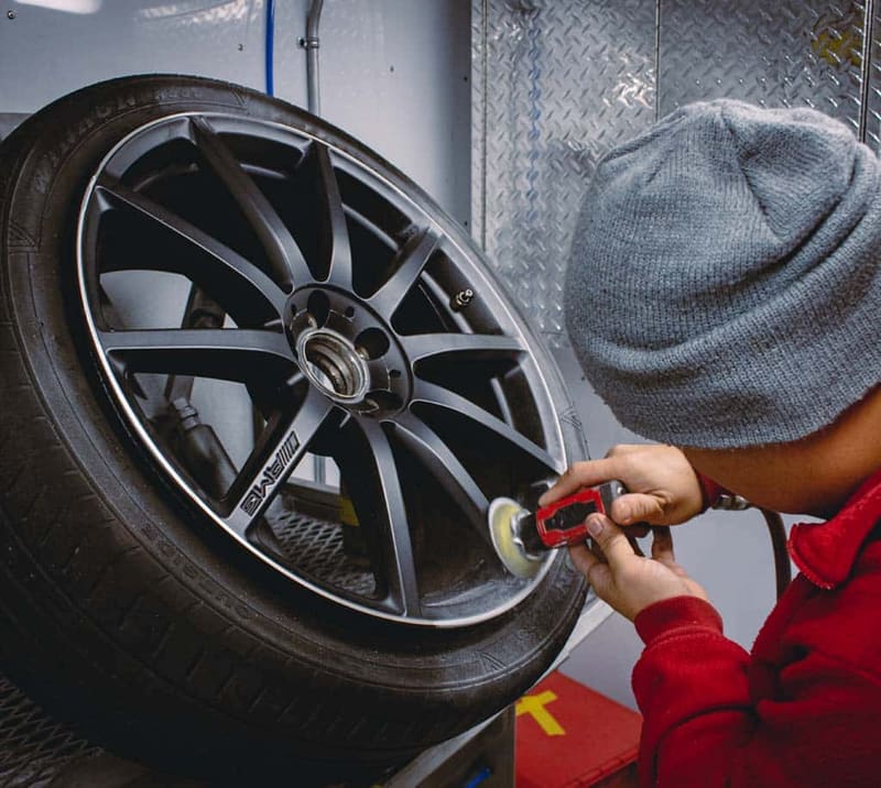 A person in a red jacket running a small orange machine polisher across the face of a dark grey alloy wheel indoors, diamond-plate wall behind