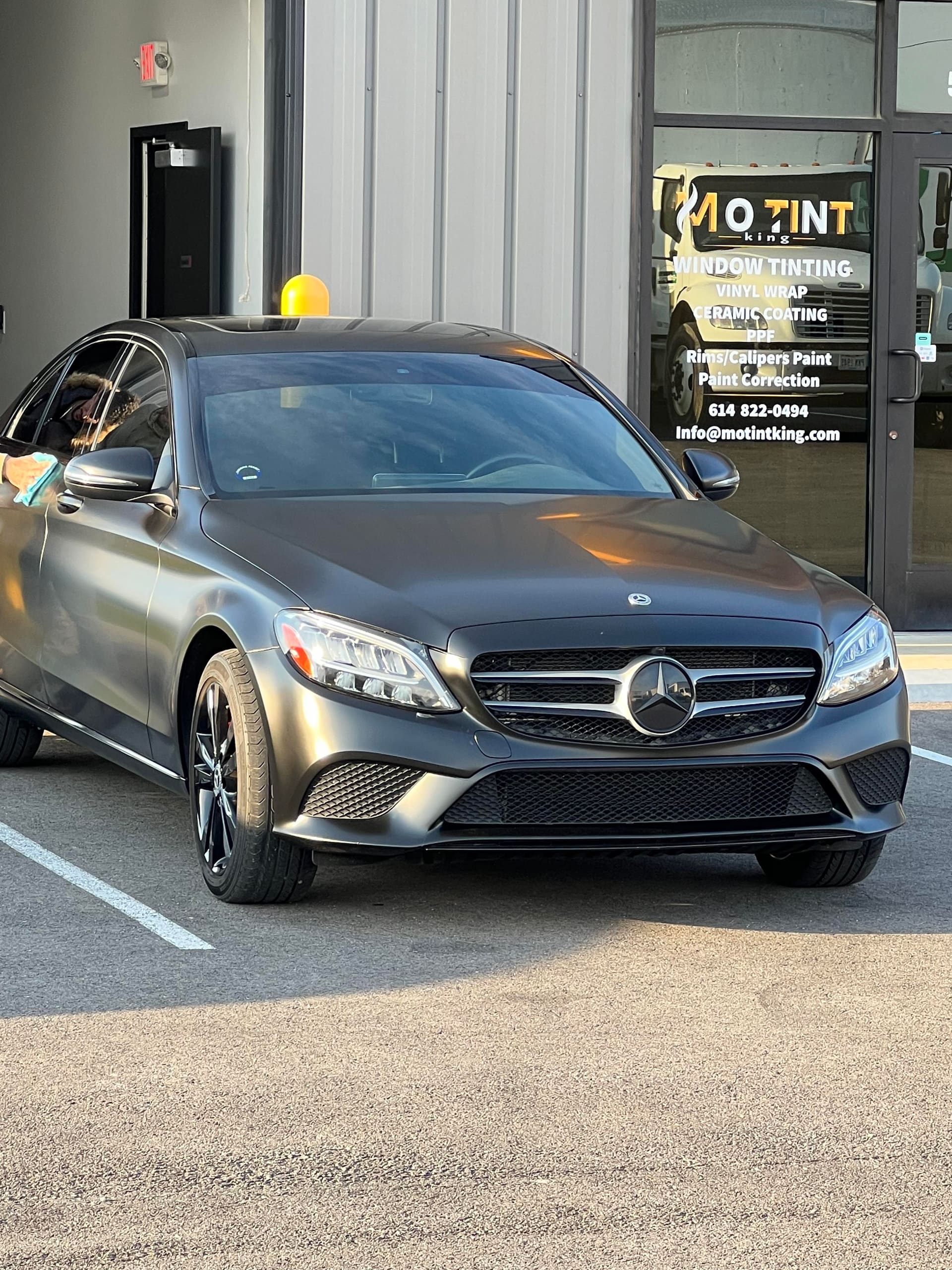 An installer's arm working a blue squeegee across the film on a grey Mercedes C-Class front door window, the MO TINT KING sign behind