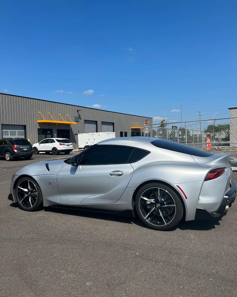 Silver Toyota GR Supra in side profile with dark tinted glass, parked in front of the shop's roller bay doors and yellow awnings