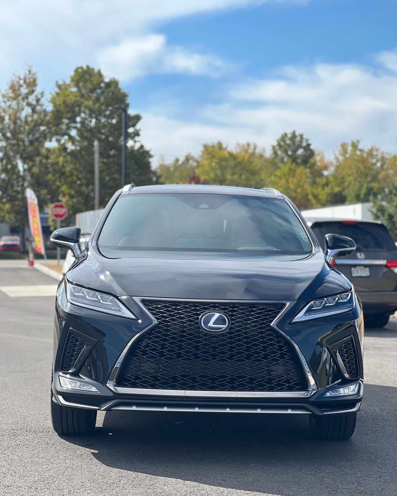 Head-on front view of a black Lexus RX F Sport showing the windshield and tinted glass, autumn trees behind