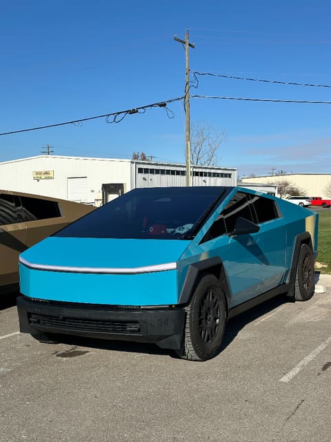 Tesla Cybertruck in a gloss cyan wrap parked outside the shop under a blue sky, a second bronze-wrapped Cybertruck partly visible behind